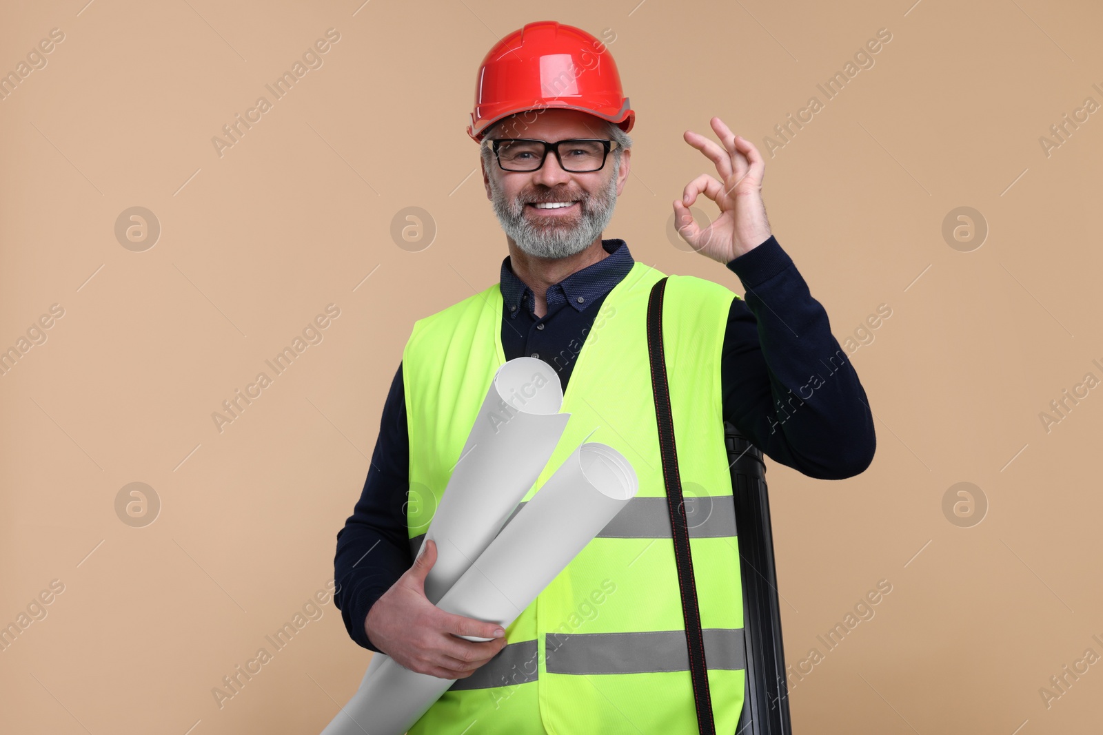 Photo of Architect in hard hat holding drafts and showing ok gesture on beige background