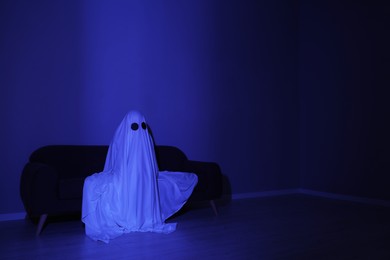 Photo of Creepy ghost. Woman covered with sheet sitting on sofa in blue light, space for text