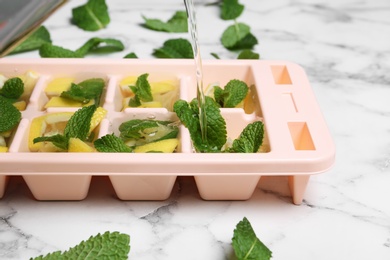 Photo of Pouring water into ice cube tray with mint and lemon on marble table