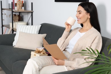 Photo of Happy woman reading greeting card while drinking coffee on sofa in living room