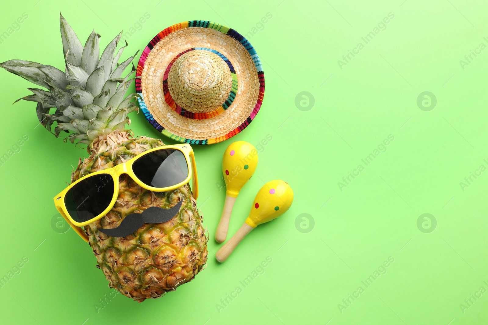 Photo of Mexican sombrero hat, pineapple with sunglasses, fake mustache and maracas on green background, flat lay. Space for text