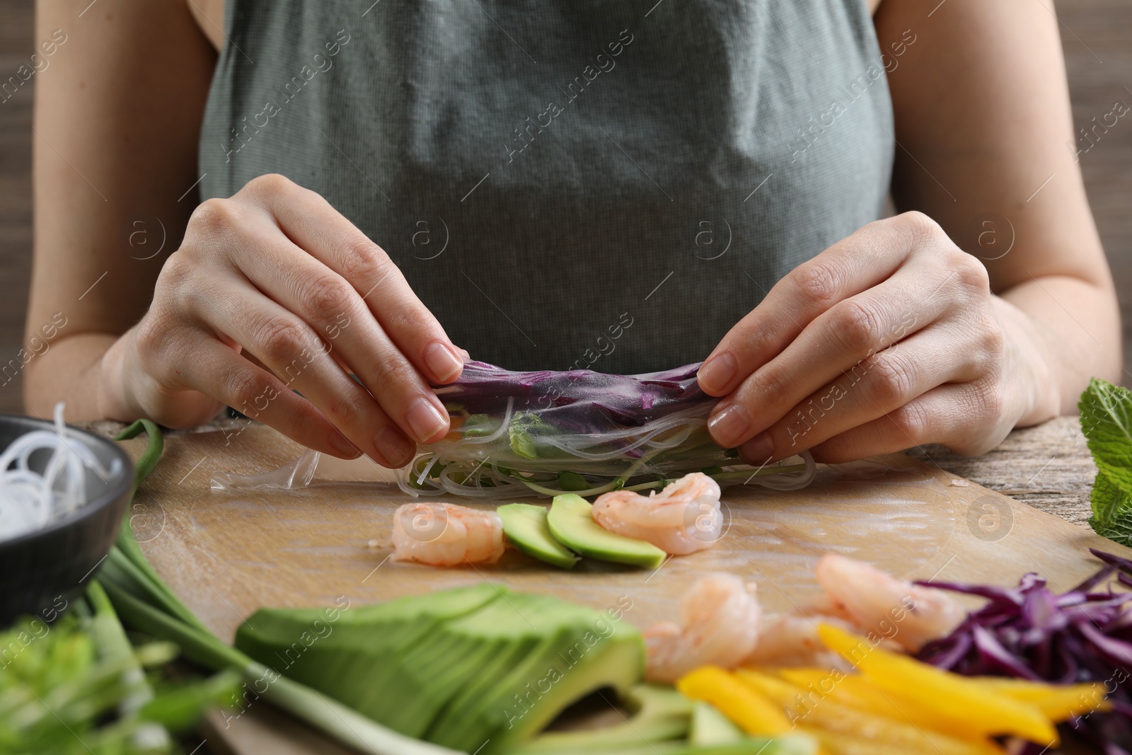 Photo of Woman wrapping spring roll at table with products, closeup