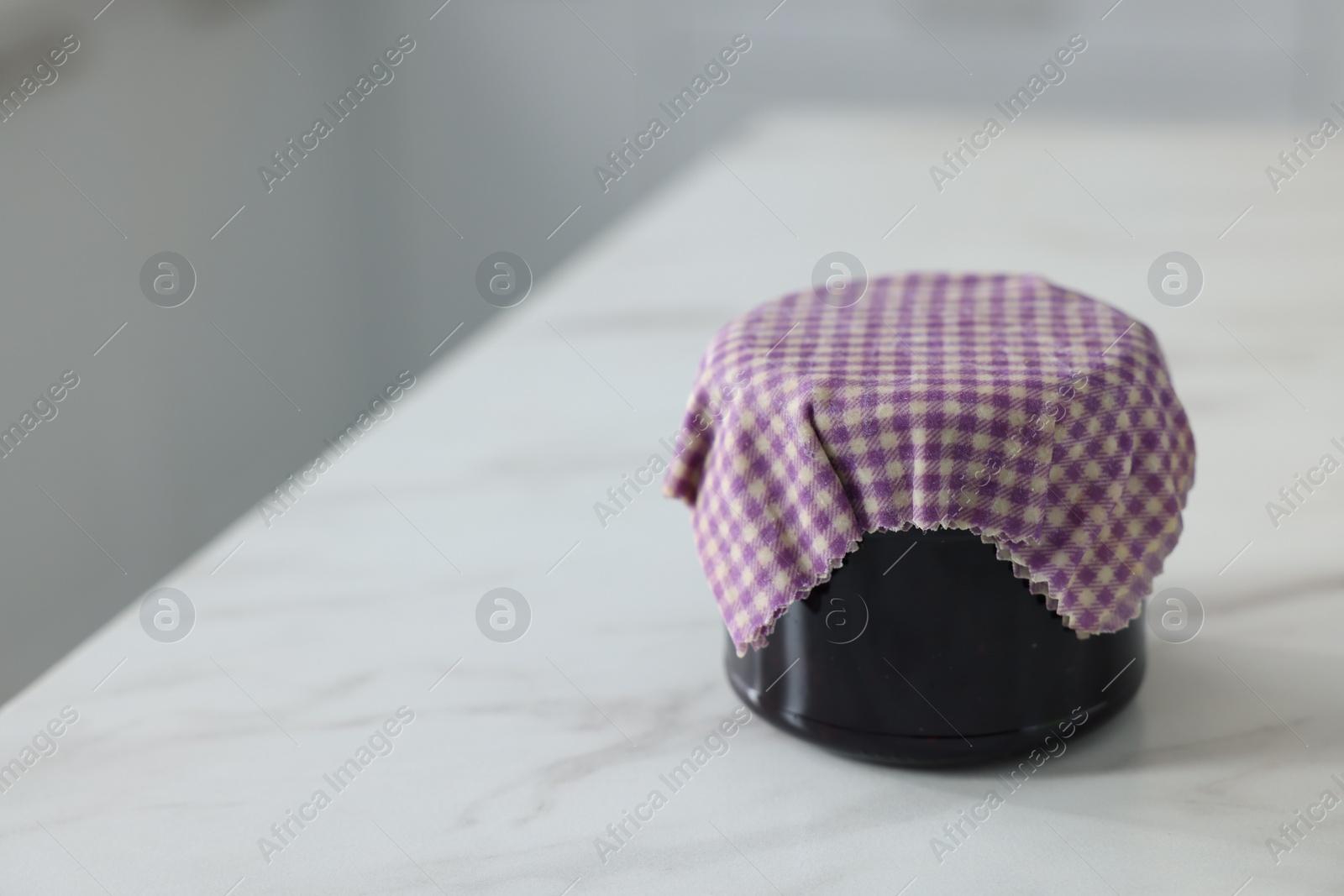 Photo of Jar of jam covered with beeswax food wrap on white table, closeup. Space for text