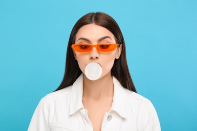 Photo of Beautiful woman in sunglasses blowing bubble gum on light blue background