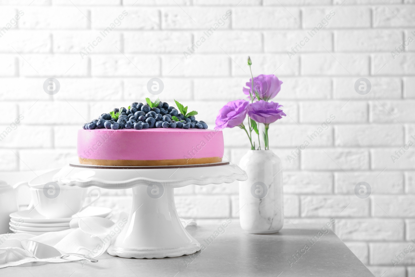 Photo of Stand with tasty blueberry cake and flowers on grey table against white brick wall. Space for text