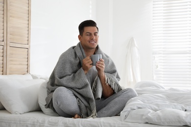 Man covered with warm grey plaid enjoying hot morning drink in bedroom