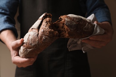 Photo of Man breaking loaf of fresh bread on dark background, closeup