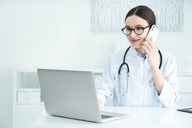 Photo of Young female doctor talking on phone at table in office