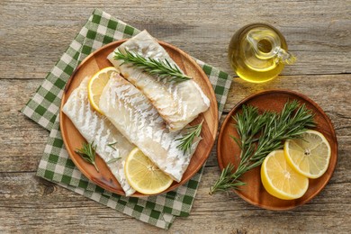 Photo of Fresh raw cod fillets with rosemary, lemon and oil on wooden table, flat lay