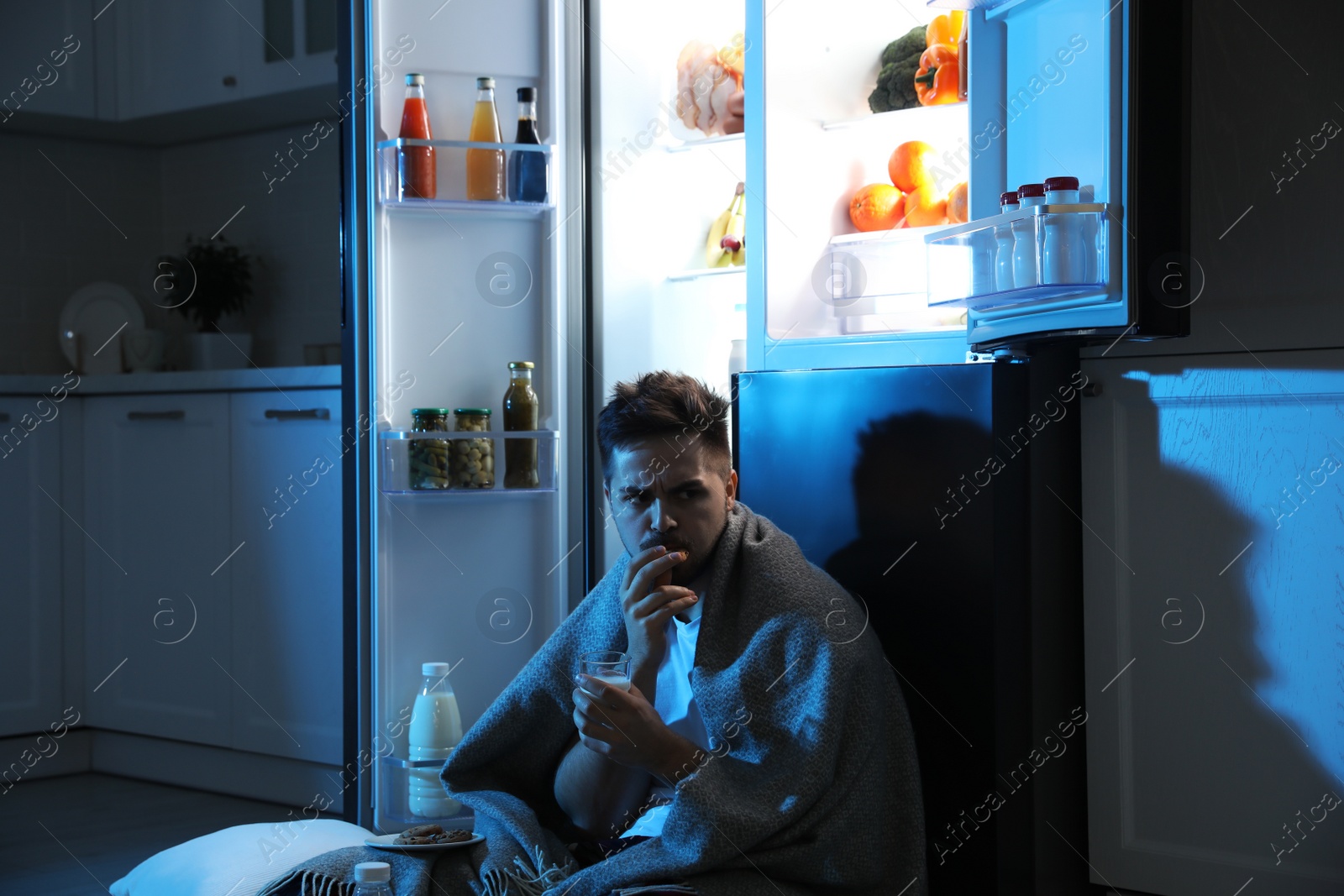 Photo of Young man eating cookies near open refrigerator in kitchen at night
