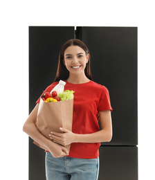 Young woman with bag of groceries near refrigerator on white background