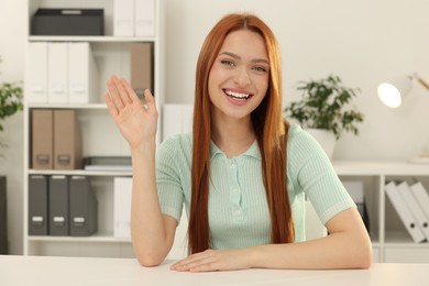 Photo of Young woman waving hello during video chat at table in office, view from web camera