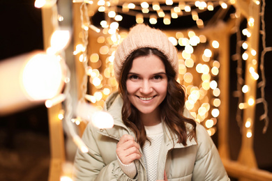 Beautiful young woman near festive lights outdoors. Winter vacation