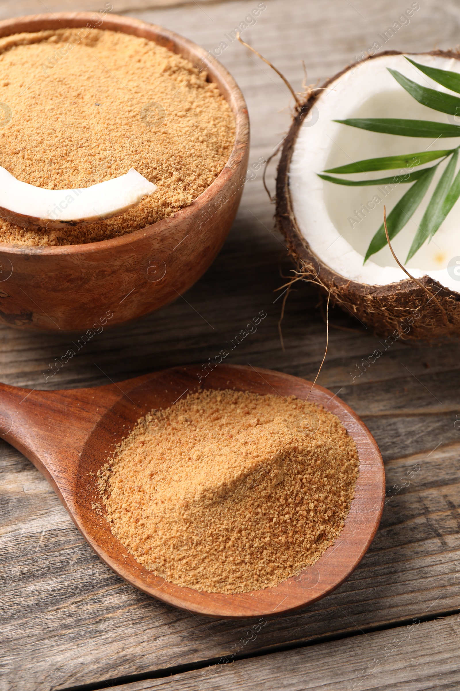 Photo of Coconut sugar in bowl, spoon, fruit and palm leaves on wooden table, closeup