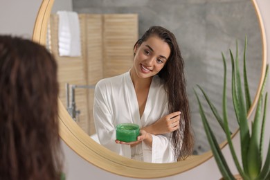 Young woman applying aloe hair mask near mirror in bathroom