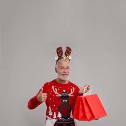 Photo of Happy senior man in Christmas sweater and reindeer headband with shopping bags showing thumbs up on grey background