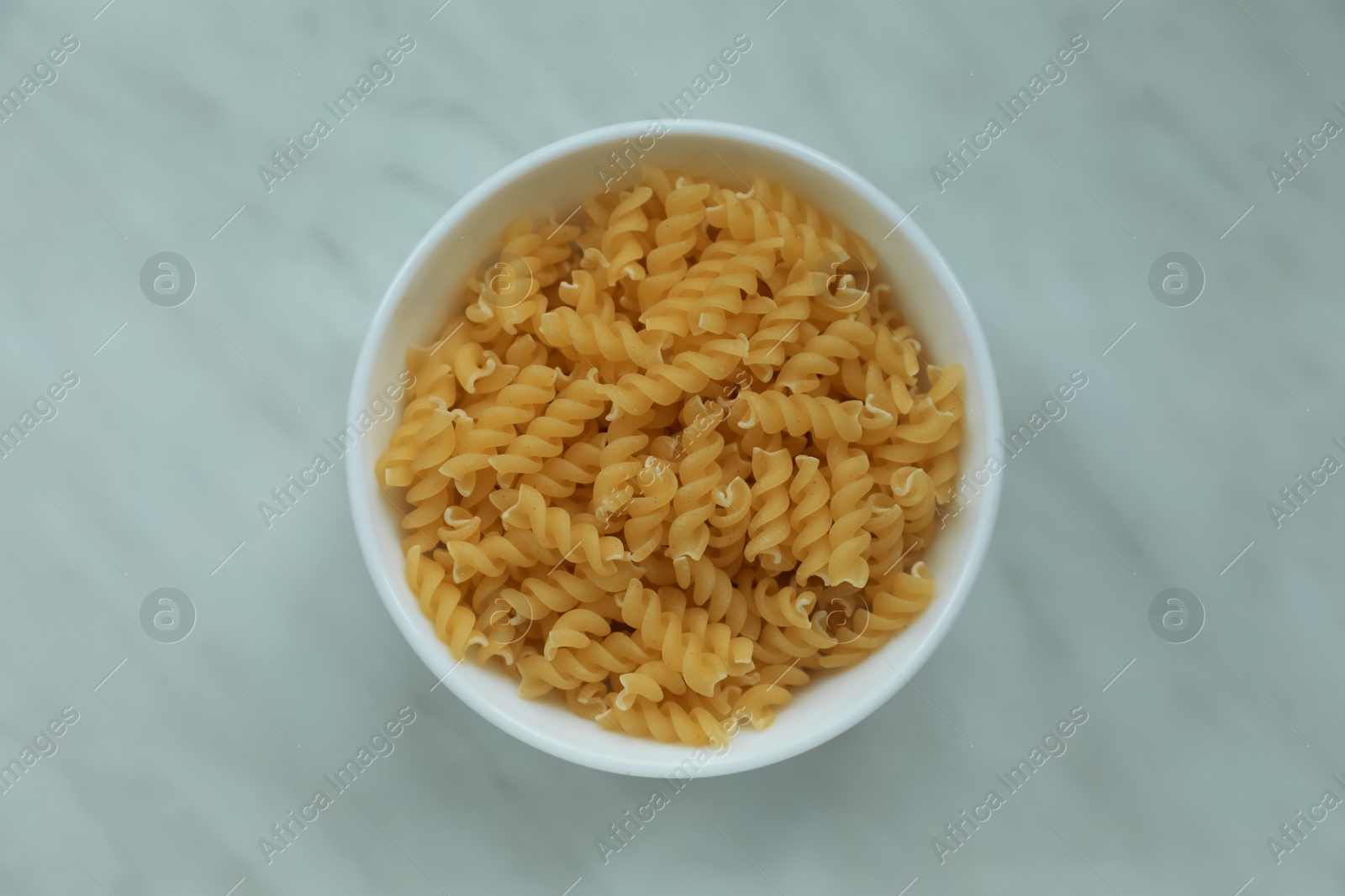 Photo of Bowl with uncooked fusilli pasta on white marble table, top view