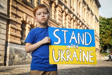Photo of Sad boy holding poster in colors of national flag and words Stand with Ukraine on city street