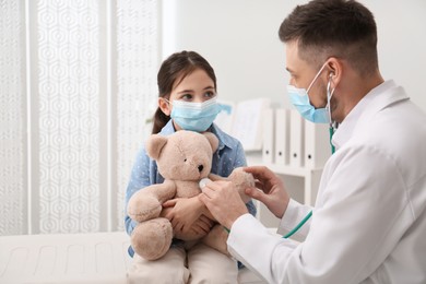 Photo of Pediatrician playing with little girl during visit in hospital. Doctor and patient wearing protective masks