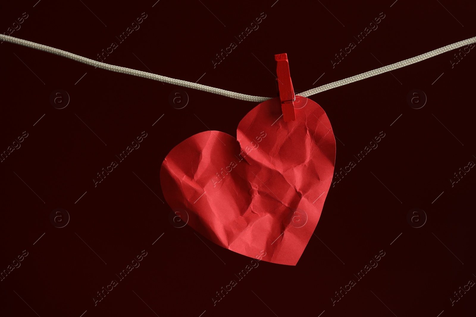 Photo of Crumpled red paper heart on rope against burgundy background. Broken heart