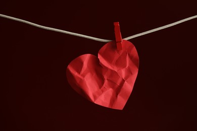 Crumpled red paper heart on rope against burgundy background. Broken heart