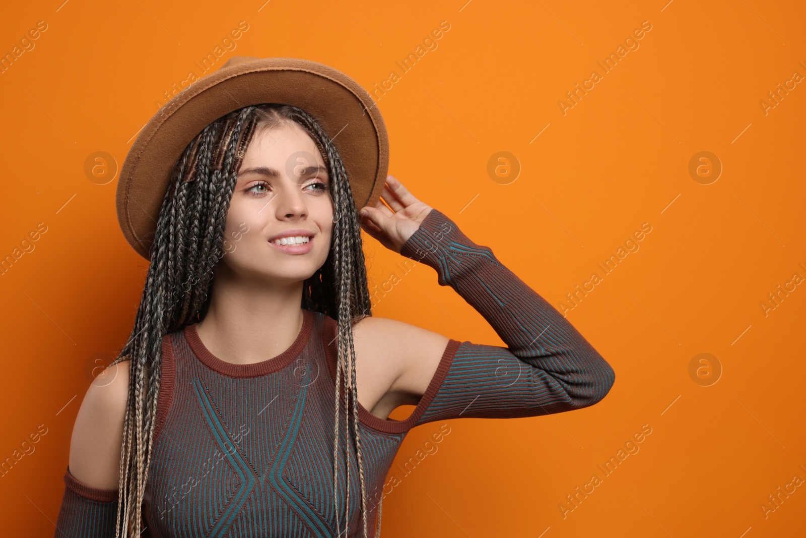 Photo of Beautiful woman with long african braids and hat on orange background, space for text
