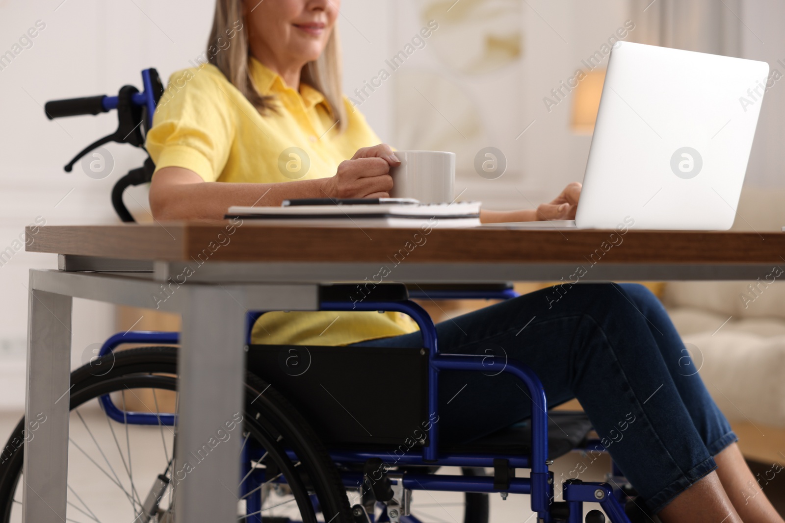 Photo of Woman in wheelchair with cup of drink using laptop at home, closeup