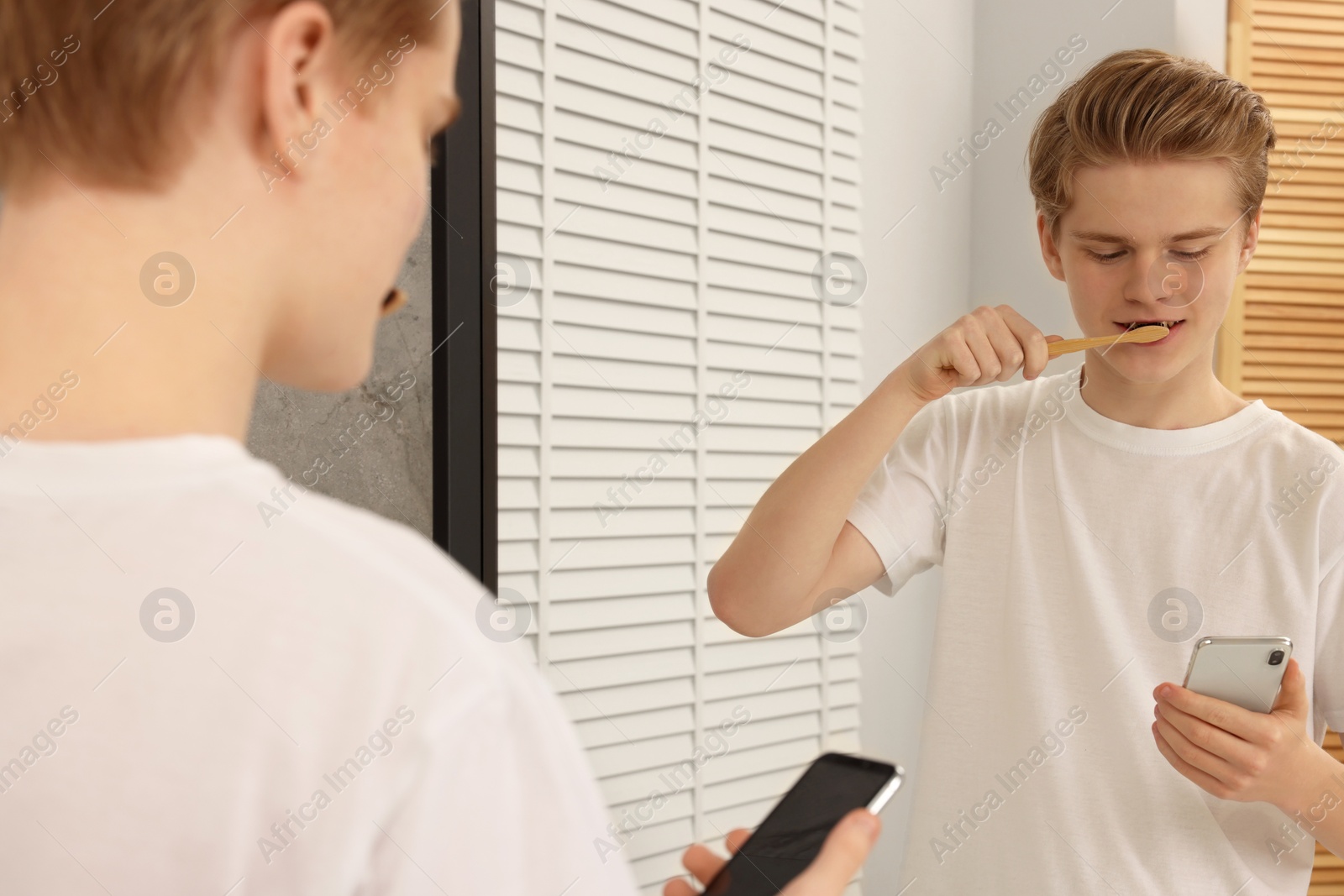 Photo of Teenage boy using smartphone while brushing teeth in bathroom. Internet addiction