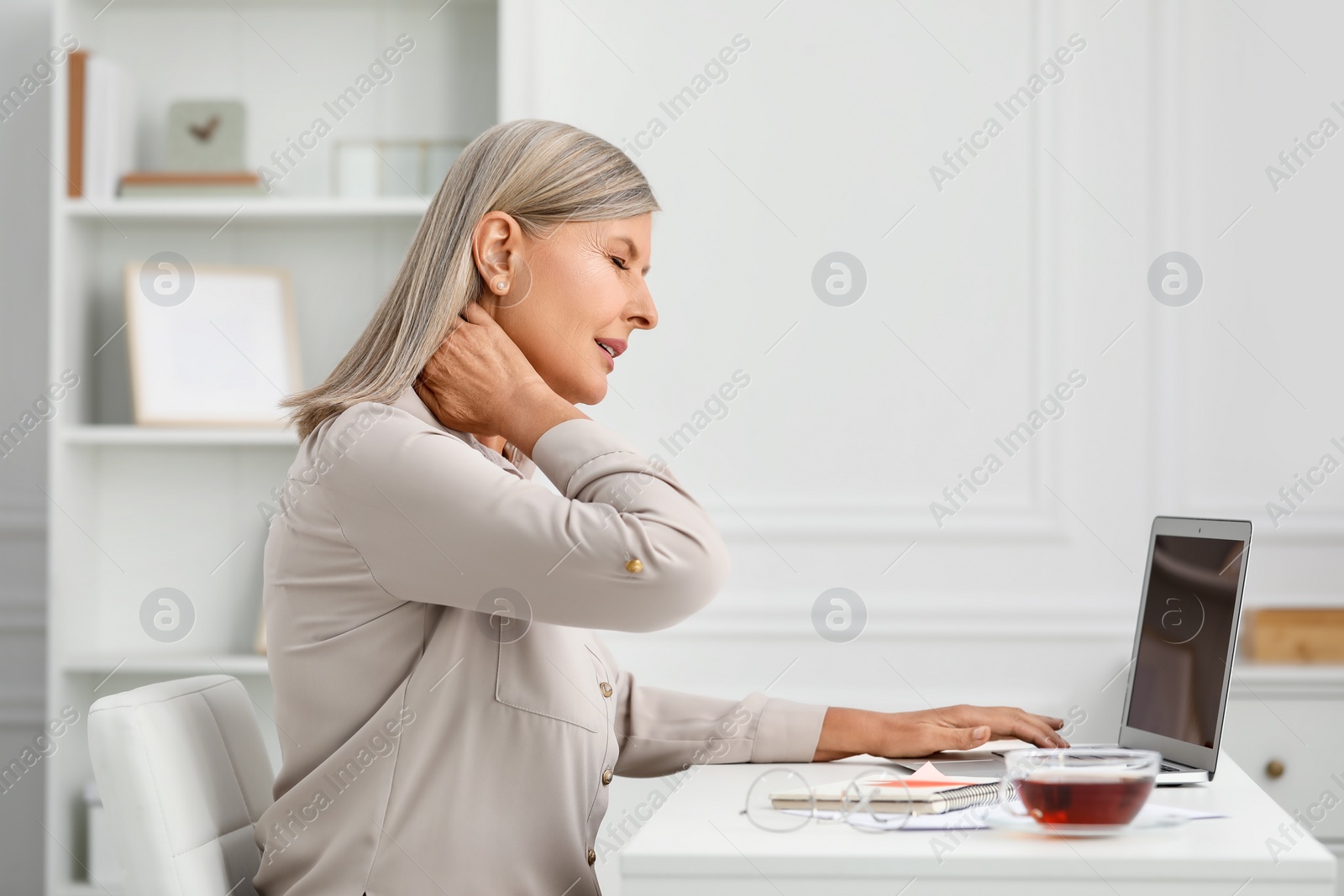 Photo of Woman suffering from neck pain at workplace in room