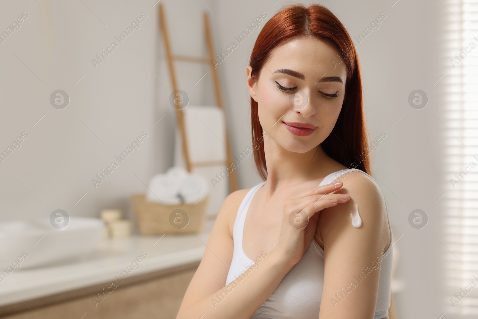 Photo of Beautiful young woman applying body cream onto shoulder in bathroom, space for text