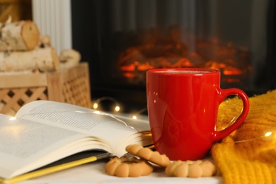 Photo of Cup of drink, book and cookies on table near fireplace