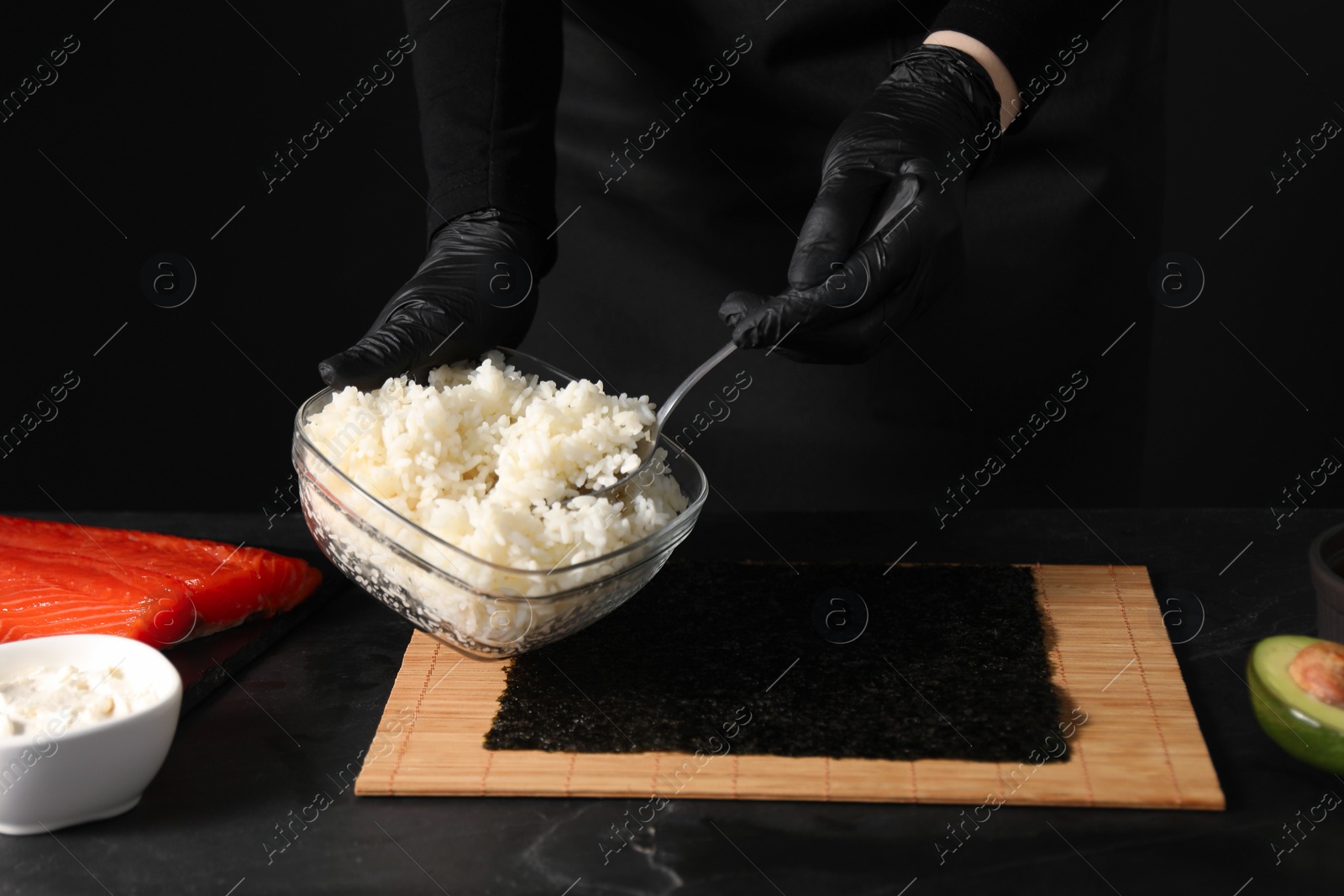 Photo of Chef in gloves taking cooked rice for sushi with spoon at dark table, closeup