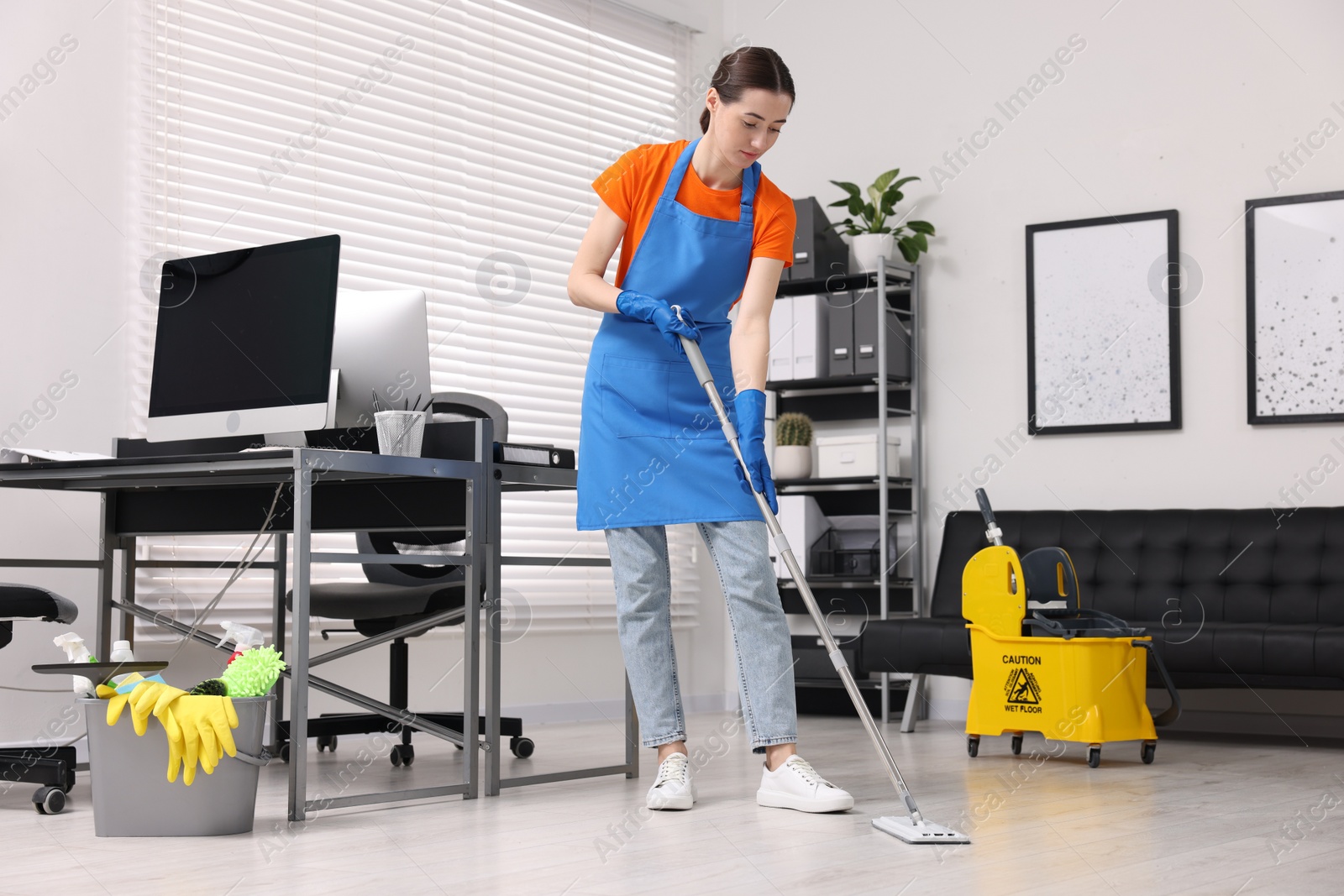 Photo of Cleaning service worker washing floor with mop. Bucket with supplies and wet floor sign in office