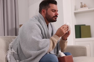 Photo of Sick man wrapped in blanket with tissues on sofa at home. Cold symptoms