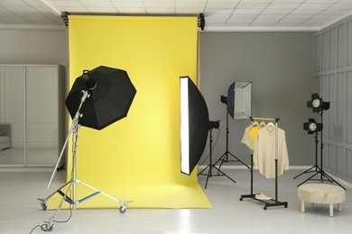 Photo of Yellow photo background, rack with clothes and professional lighting equipment in modern studio