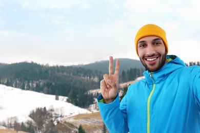 Image of Smiling young man taking selfie and showing peace sign in mountains