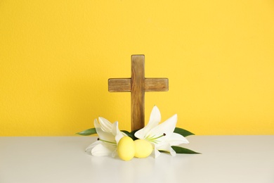 Photo of Wooden cross, Easter eggs and blossom lilies on table against color background