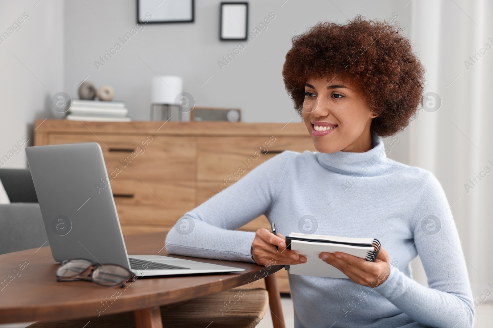 Photo of Beautiful young woman using laptop and writing in notebook at wooden coffee table in room