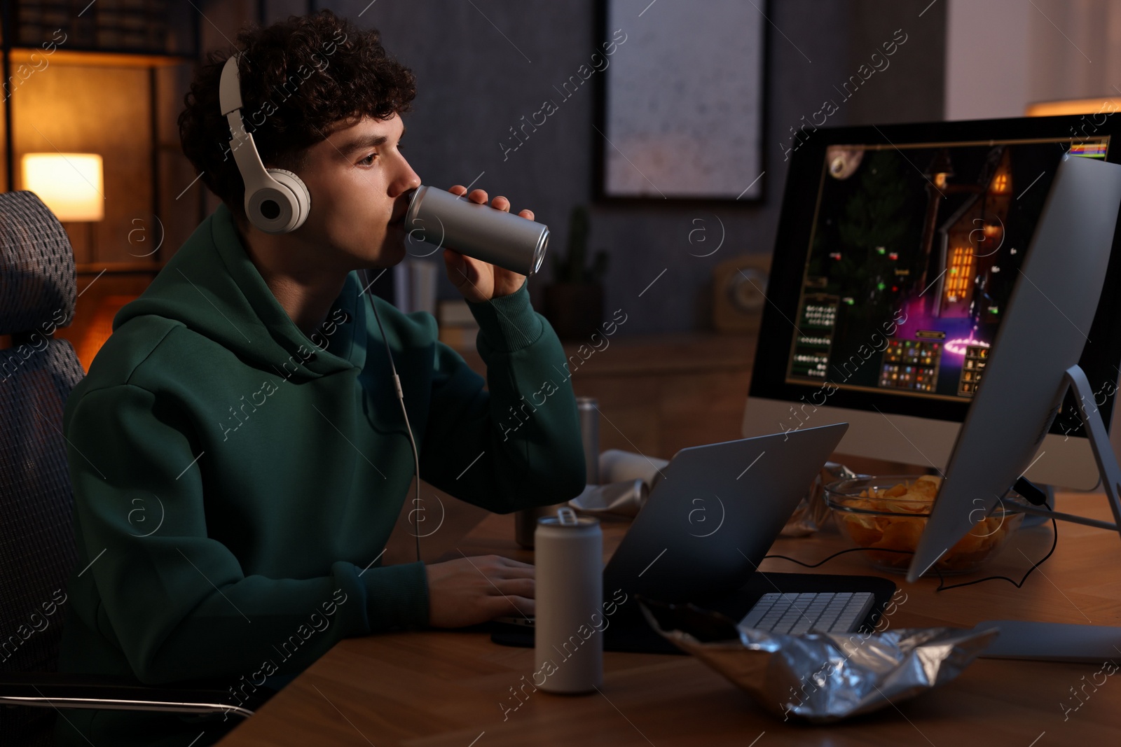 Photo of Young man with energy drink and headphones playing video game at wooden desk indoors