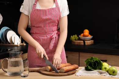 Woman in striped apron cutting baguette on wooden table at kitchen, closeup