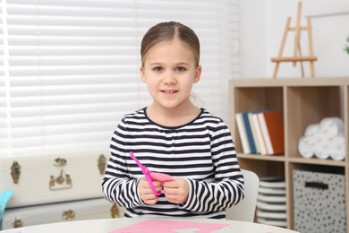 Cute little girl with scissors and pink paper at desk in room. Home workplace