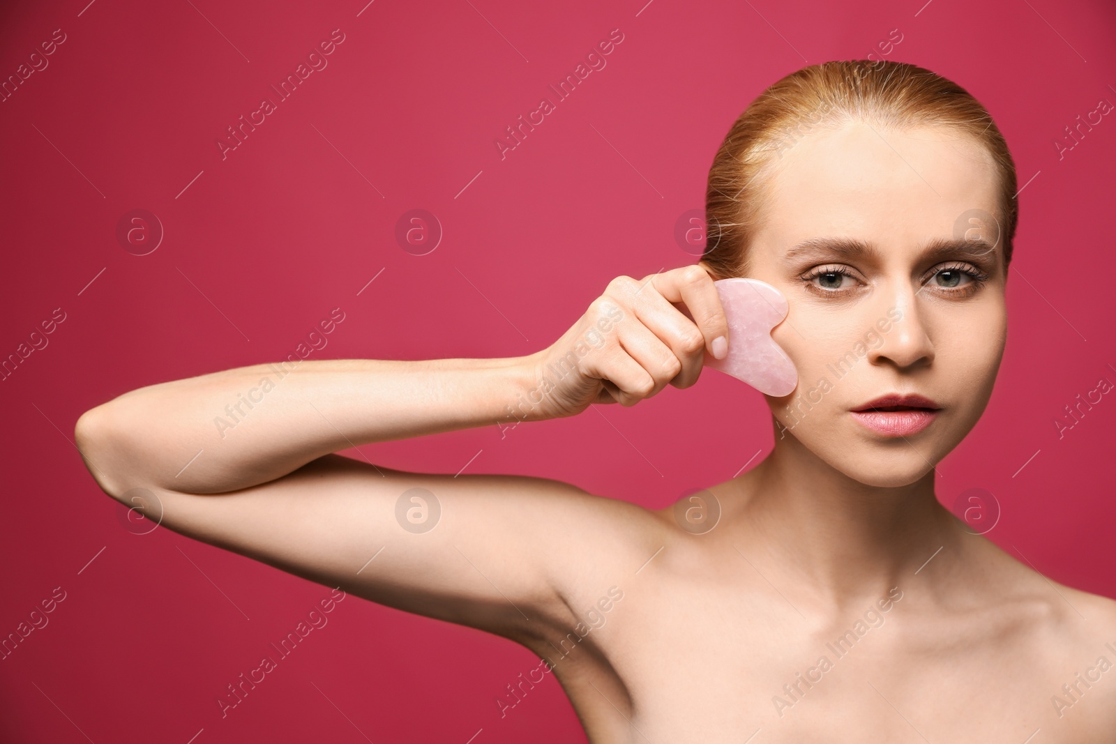 Photo of Beautiful young woman doing facial massage with gua sha tool on pink background