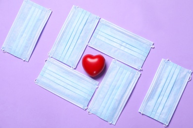 Red decorative heart among medical masks on violet background, flat lay