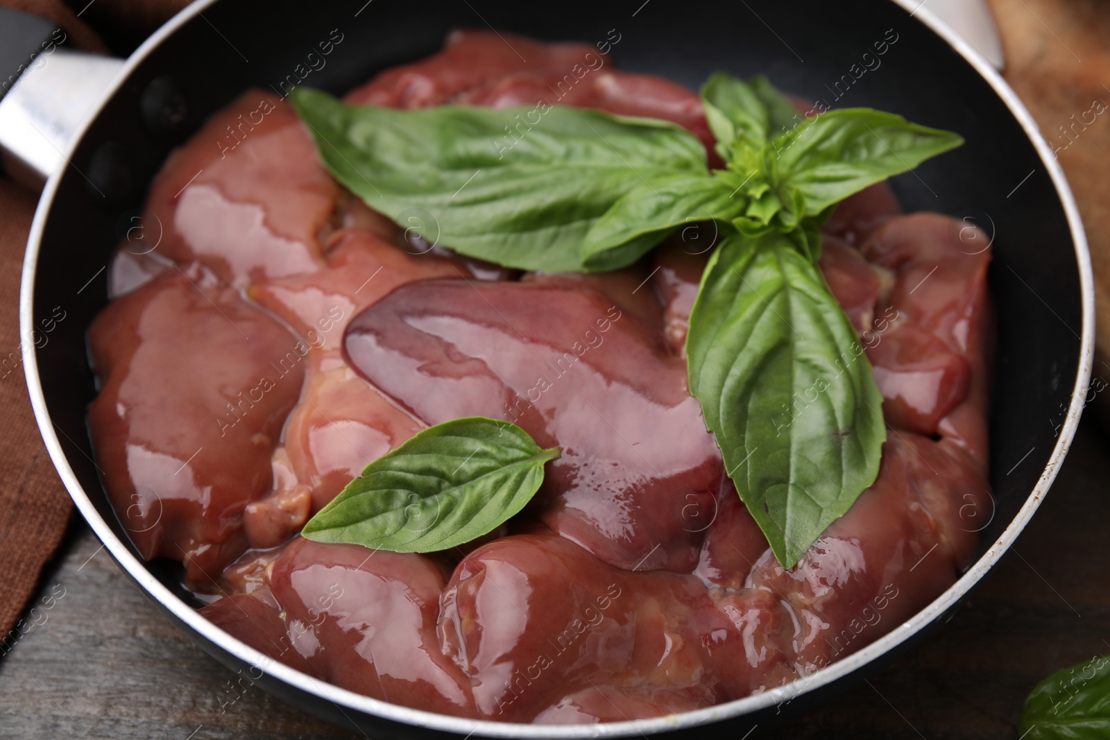 Photo of Raw chicken liver with basil in frying pan on wooden table, closeup