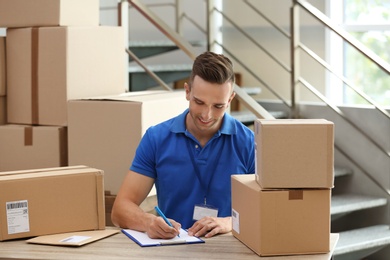 Young courier working with papers among parcels at table in delivery department