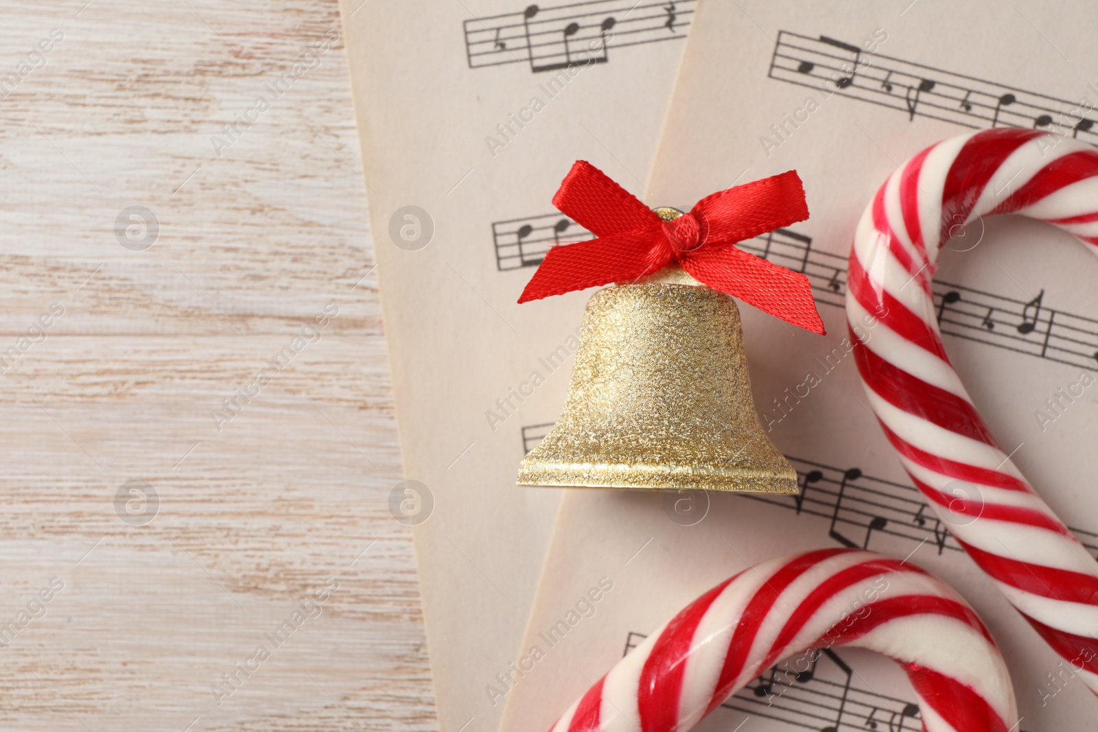 Photo of Golden shiny bell with red bow, music sheets and candy canes on wooden table, flat lay. Space for text. Christmas decoration