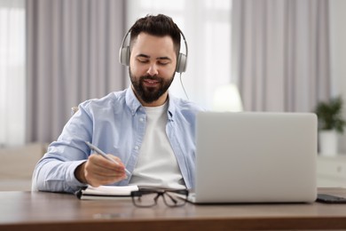 Photo of Young man in headphones writing down notes during webinar at table in room