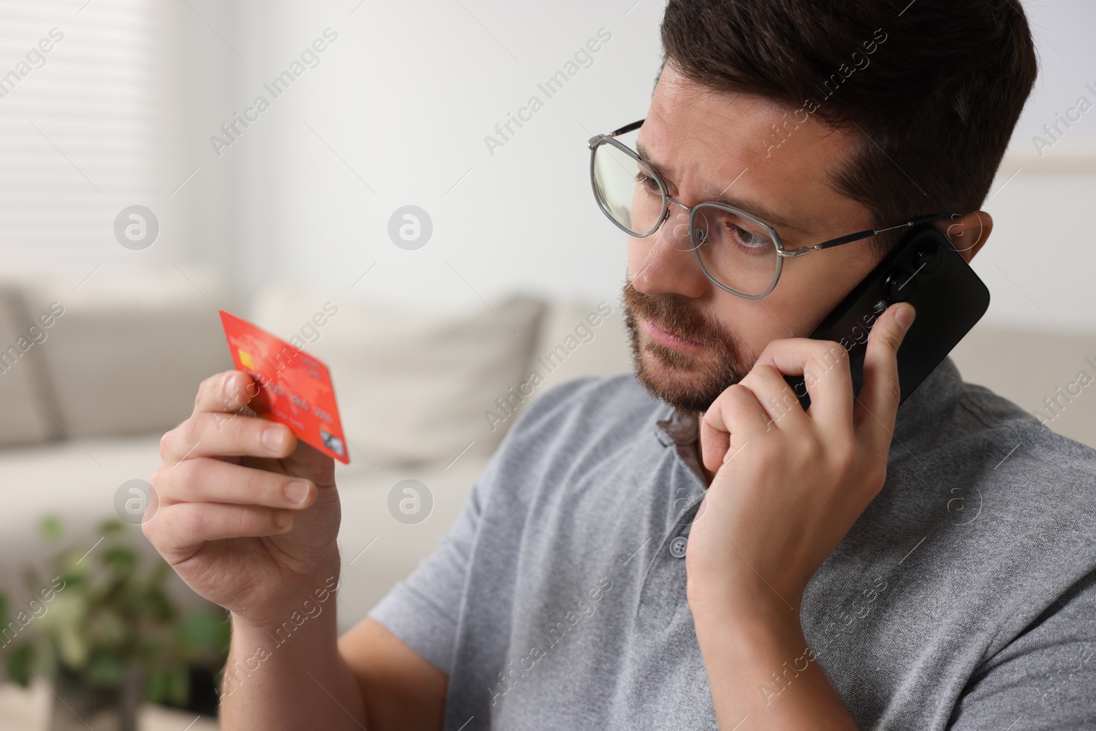 Photo of Upset man with credit card talking on smartphone at home. Be careful - fraud