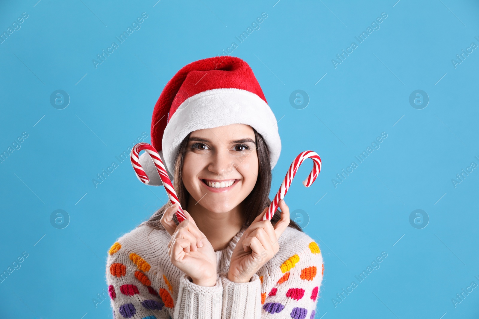 Photo of Pretty woman in Santa hat and festive sweater holding candy canes on light blue background