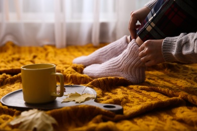 Photo of Woman relaxing with cup of hot winter drink on knitted plaid indoors, closeup. Cozy season